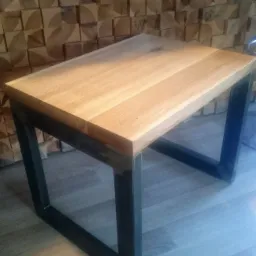 Stolik kawowy z blatem z jasnego drewna i czarnymi, metalowymi nogami, na tle ściany z drewnianych kostek.