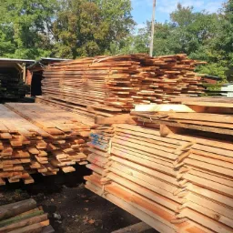 Stosy świeżo przetartego drewna ułożone na zewnątrz, widoczne oznaczenia kolorową kredą na niektórych deskach, w tle zieleń drzew i słup oświetleniowy.