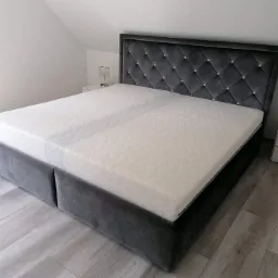 Szare tapicerowane łóżko z pikowanym zagłówkiem zdobionym kryształkami, ustawione w jasnym pokoju na drewnianej podłodze, obok biała szafka nocna z lampką.