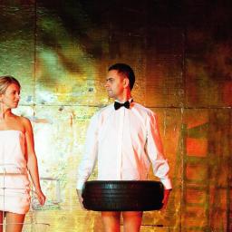 Fotograf ślubny - Para w teatralnych kostiumach na tle abstrakcyjnej scenografii: kobieta w białej sukience z drucianą klatką, mężczyzna w koszuli i muszce z oponą samochodową, w tle drabina z modelem Krzywej Wieży...