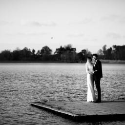 Fotograf ślubny - Czarno-białe zdjęcie pary młodej stojącej na drewnianym pomoście nad jeziorem, w tle las i pochmurne niebo.