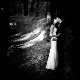 Fotograf ślubny - Czarno-białe zdjęcie całującej się pary młodej w lesie, panna młoda w koronkowej sukni, pan młody w smokingu, ścieżka usłana liśćmi.