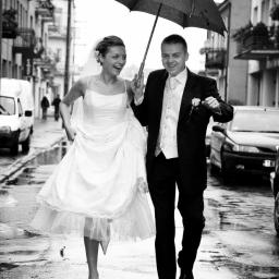 Fotograf ślubny - Czarno-białe zdjęcie uśmiechniętej pary młodej biegnącej po mokrej ulicy pod parasolem, z widocznymi budynkami i samochodami w tle.