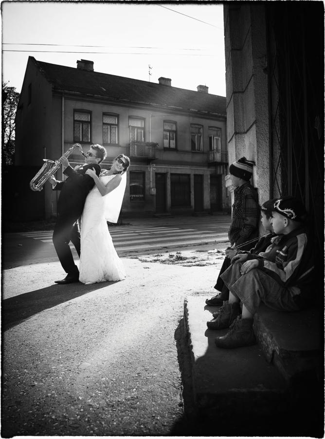 Czarno-białe zdjęcie pary młodej na ulicy: pan młody gra na saksofonie, panna młoda uśmiecha się w okularach słonecznych, w tle budynek i obserwujący ich chłopcy.