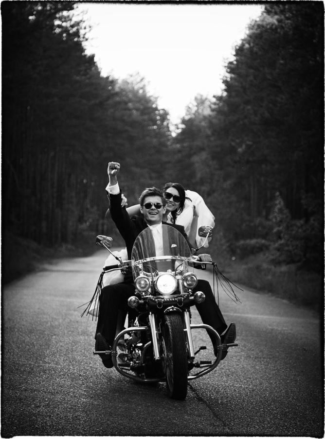 Czarno-białe zdjęcie nowożeńców na motocyklu, jadących leśną drogą, pan młody z uniesioną pięścią w geście radości, panna młoda obejmuje go od tyłu.