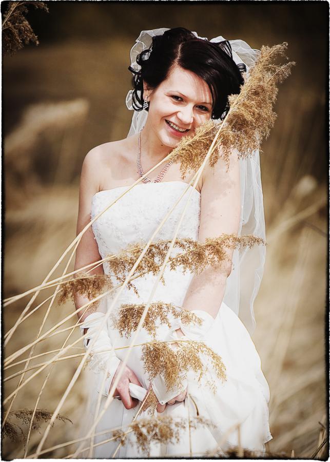 Uśmiechnięta panna młoda w białej sukni i welonie, pozująca na tle łąki z suchymi trawami, z delikatnym naszyjnikiem i kolczykami.
