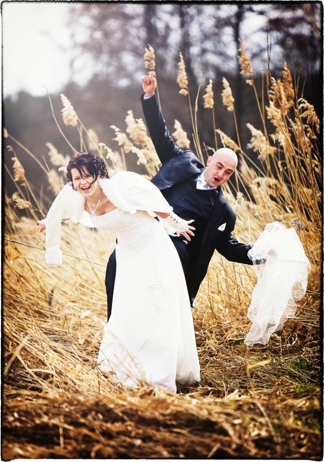 Uśmiechnięta panna młoda w białej sukni i futerku, oraz podekscytowany pan młody w czarnym garniturze, radośnie pozujący wśród wysokich traw w plenerze.