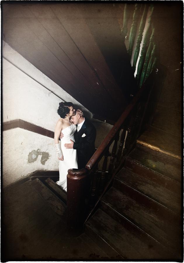 Para młoda w objęciach na drewnianych schodach z zielonymi balustradami, panna młoda w białej sukni bez ramiączek, pan młody w czarnym garniturze, ujęcie z góry w klimatycznym oświetleniu.