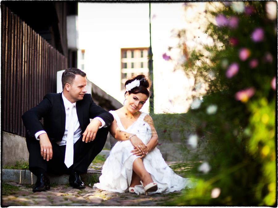 Para młoda siedzi na brukowanych schodach; on w czarnym garniturze i białym krawacie, ona w białej sukni z kwiatowym akcentem we włosach, obok kwitnące kwiaty.