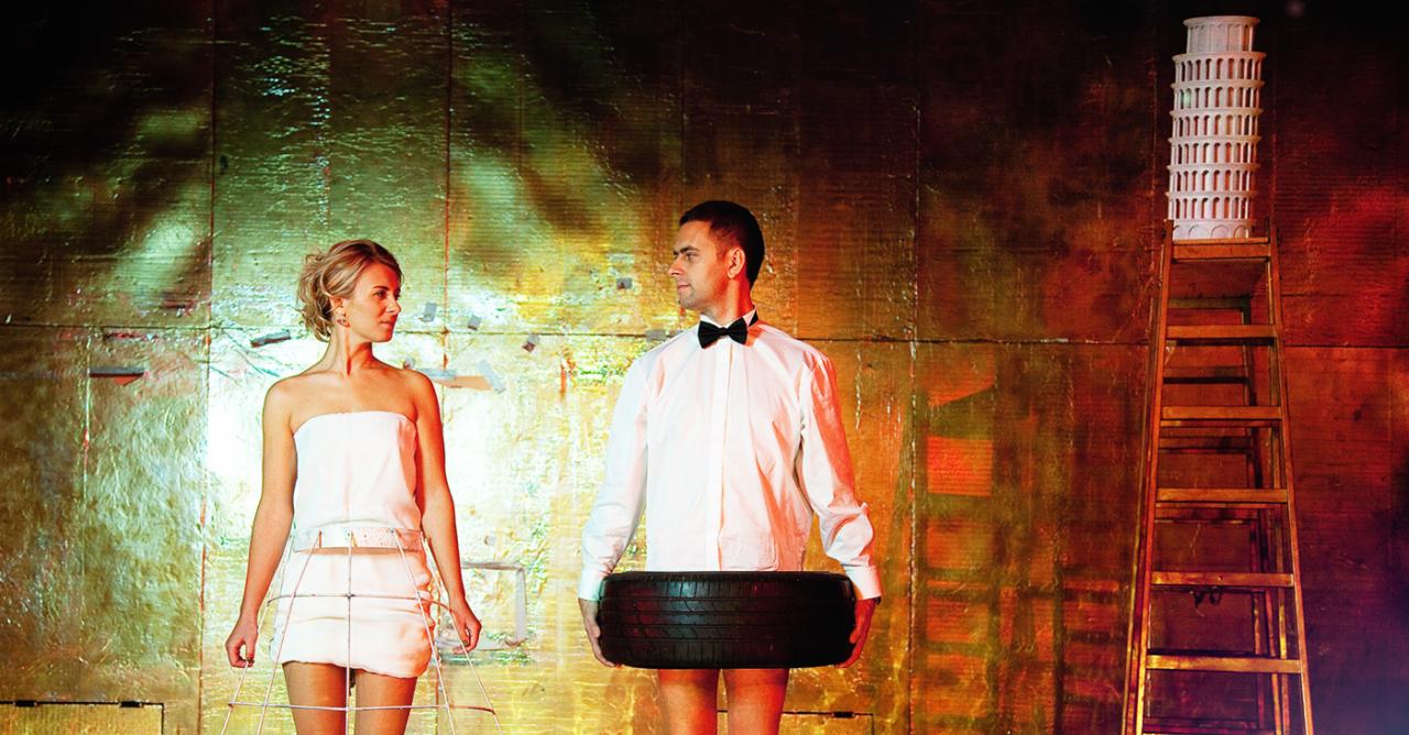 Para w teatralnych kostiumach na tle abstrakcyjnej scenografii: kobieta w białej sukience z drucianą klatką, mężczyzna w koszuli i muszce z oponą samochodową, w tle drabina z modelem Krzywej Wieży...