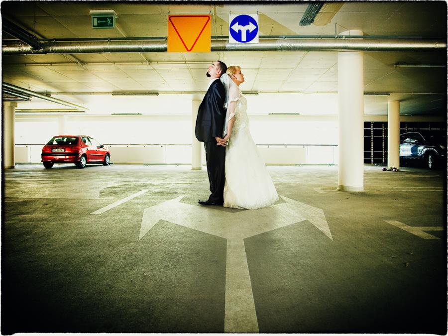 Para młoda stoi tyłem do siebie na parkingu podziemnym, pod znakami drogowymi, trzymając się za ręce; nietypowa sceneria ślubna.