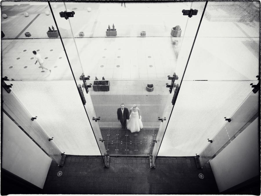 Czarno-białe zdjęcie z góry przedstawiające parę młodą stojącą na mozaikowej podłodze, widoczną przez szklaną konstrukcję z metalowymi mocowaniami, z elementami dekoracyjnymi w tle i osobą w ruchu.