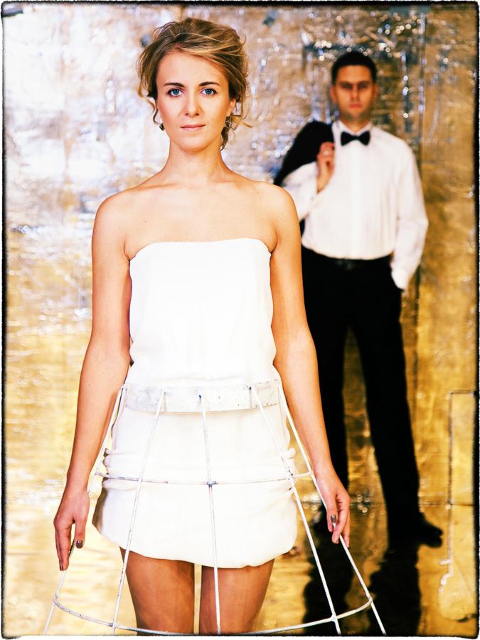 Portret kobiety w białej sukience z metalową konstrukcją, za nią mężczyzna w muszce i marynarce na ramieniu, na tle błyszczącej ściany.