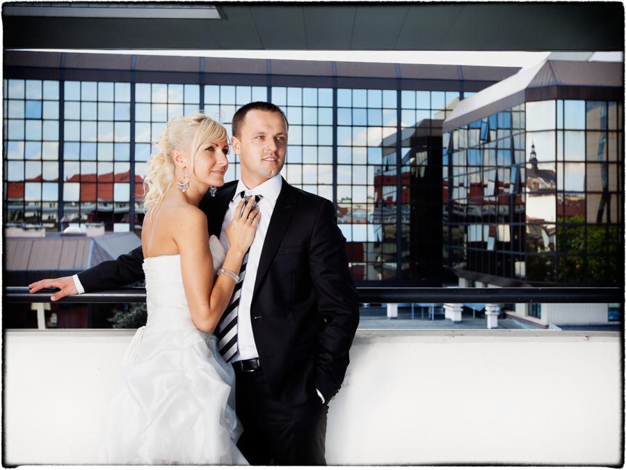 Para młoda na tle nowoczesnego budynku z lustrzanymi oknami, panna młoda w sukni bez ramiączek, pan młody w czarnym garniturze i krawacie w paski, delikatne oświetlenie.