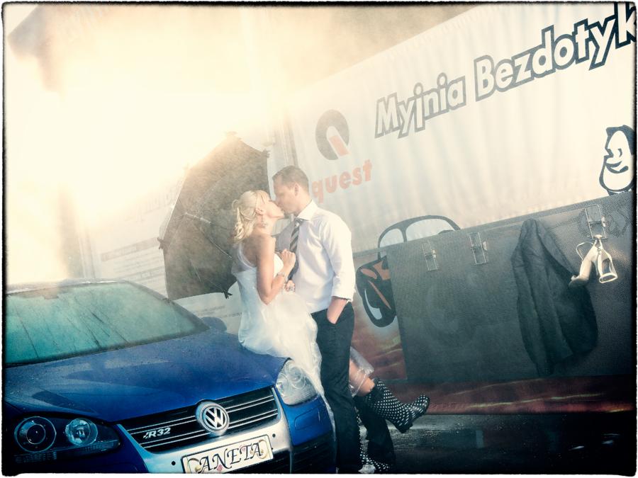 Pocałunek nowożeńców w deszczu na masce niebieskiego Volkswagena R32, z parasolem i reklamą myjni samochodowej w tle.