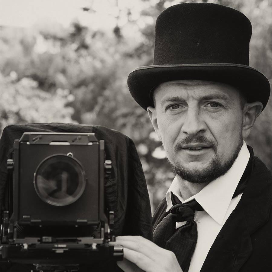 Portret fotografa w cylindrze z zabytkowym aparatem wielkoformatowym, czarno-białe ujęcie.