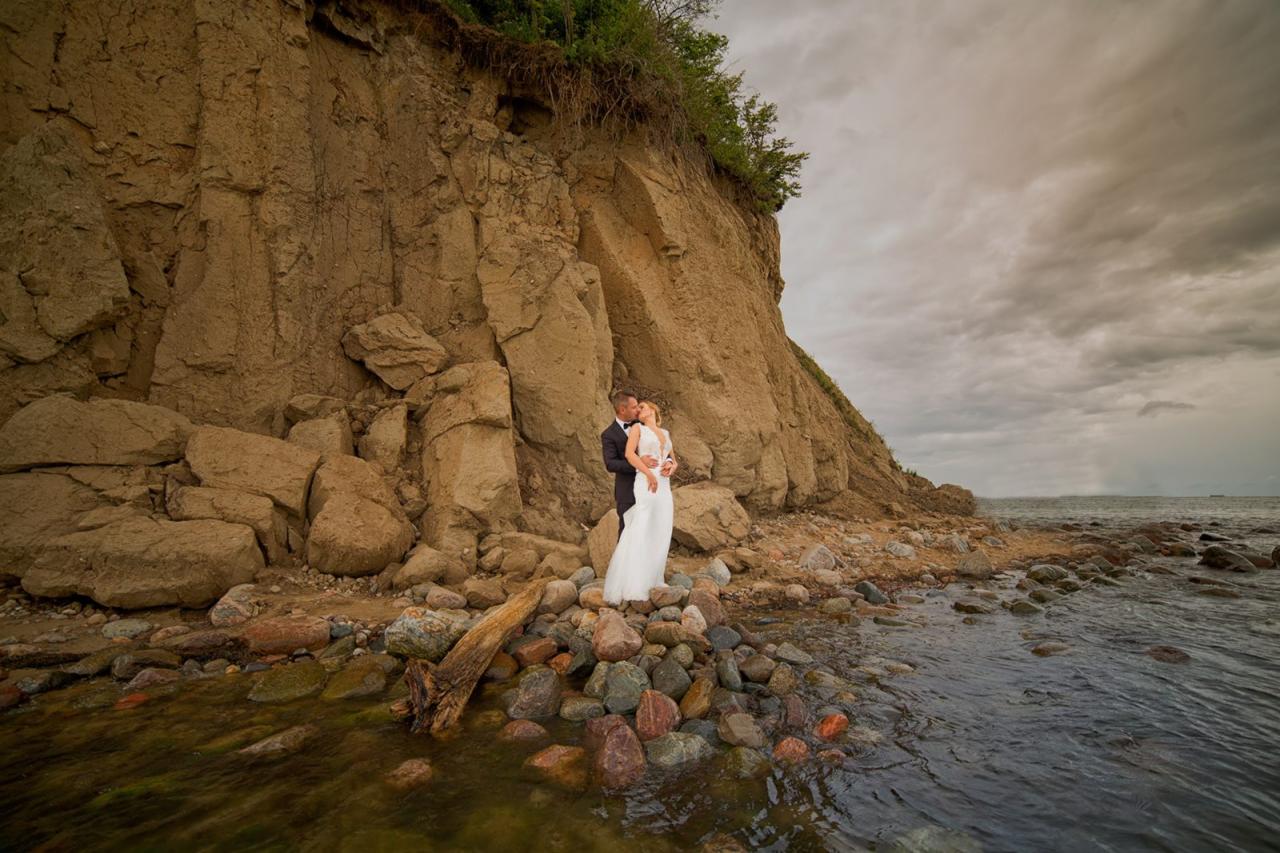 Para młoda w objęciach na kamienistej plaży u stóp klifu, w tle pochmurne niebo i morze.