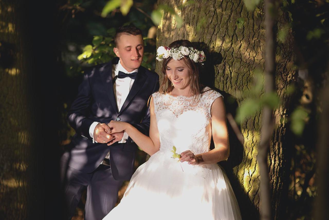 Para młoda w leśnej scenerii, panna młoda w białej sukni z koronkową górą i wiankiem, pan młody w granatowym garniturze z muchą, trzymają się za ręce opierając o korę drzewa w promieniach słońca.