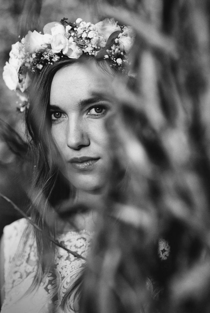 Portret kobiety w wianku z kwiatów, czarno-białe zdjęcie z ostrością na twarz, częściowo zasłoniętą przez źdźbła trawy.
