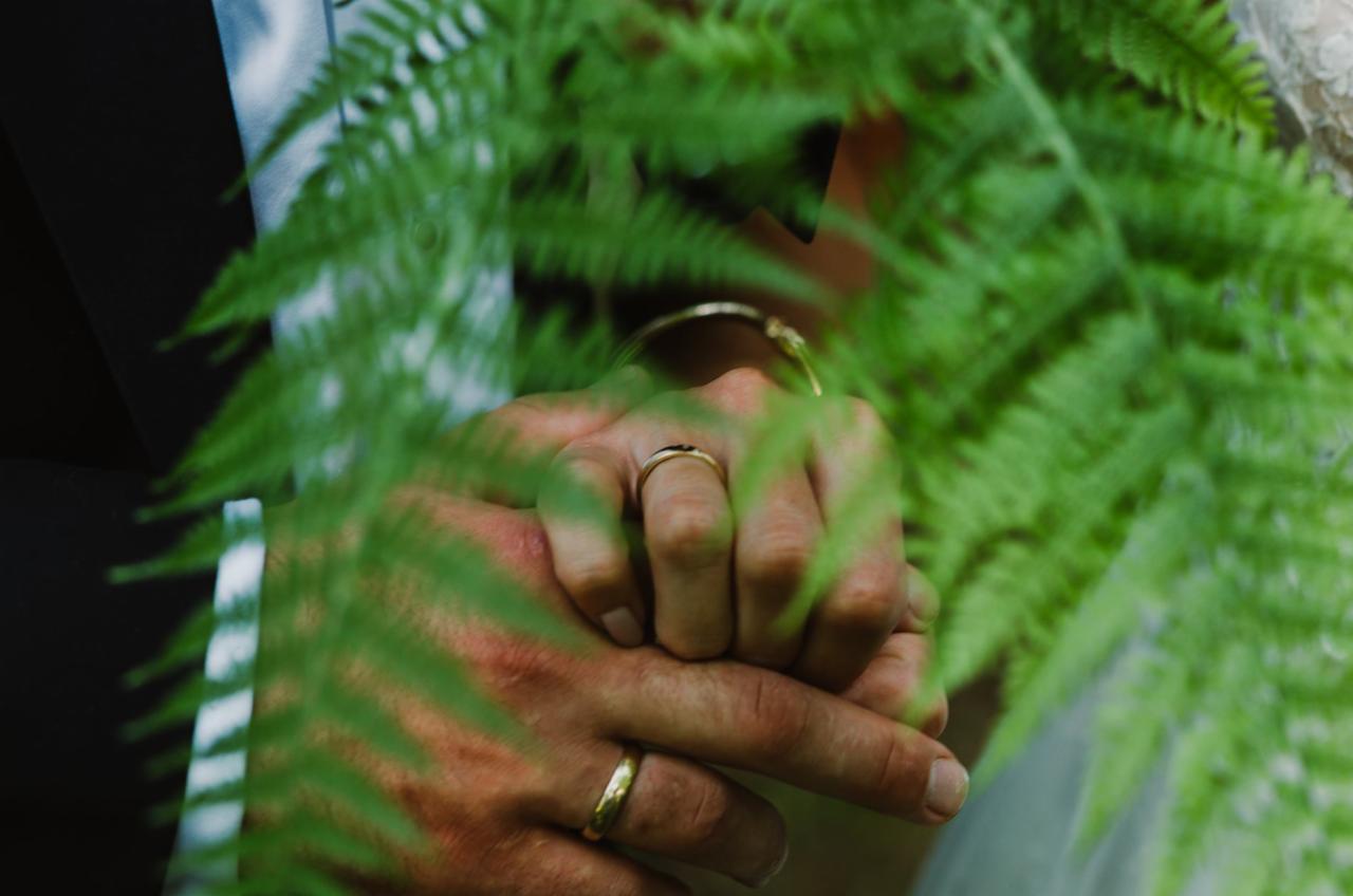 Złączone dłonie pary z obrączkami ślubnymi, częściowo zasłonięte zielonymi liśćmi paproci, w tle widoczny garnitur i suknia ślubna.