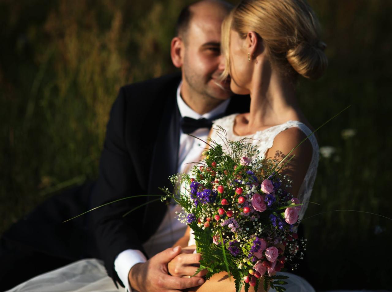 Para młoda siedząca na trawie, panna młoda trzyma bukiet z różowych i fioletowych kwiatów, pan młody w czarnym smokingu z muszką, delikatne światło słoneczne.