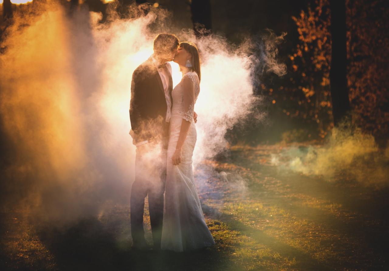Para młoda w objęciach, pocałunek w otoczeniu dymu i promieni słonecznych, plenerowa sesja ślubna.