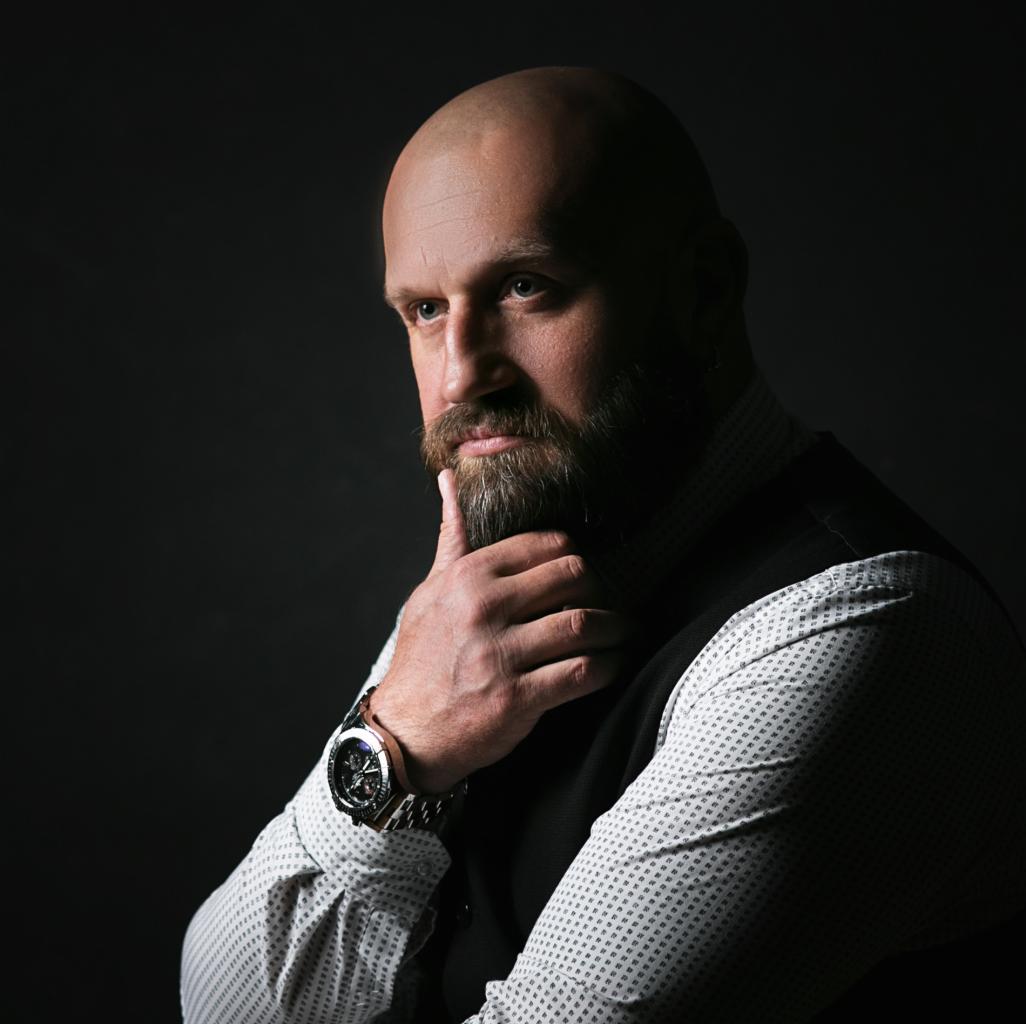 Portret brodatego mężczyzny w kamizelce, z zegarkiem na ręce, w zamyślonej pozie na ciemnym tle.