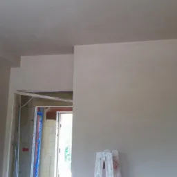 Świeżo położona, jasna, gładka warstwa na ścianach i suficie w trakcie remontu; widoczna rama drzwi i drabina w pomieszczeniu.