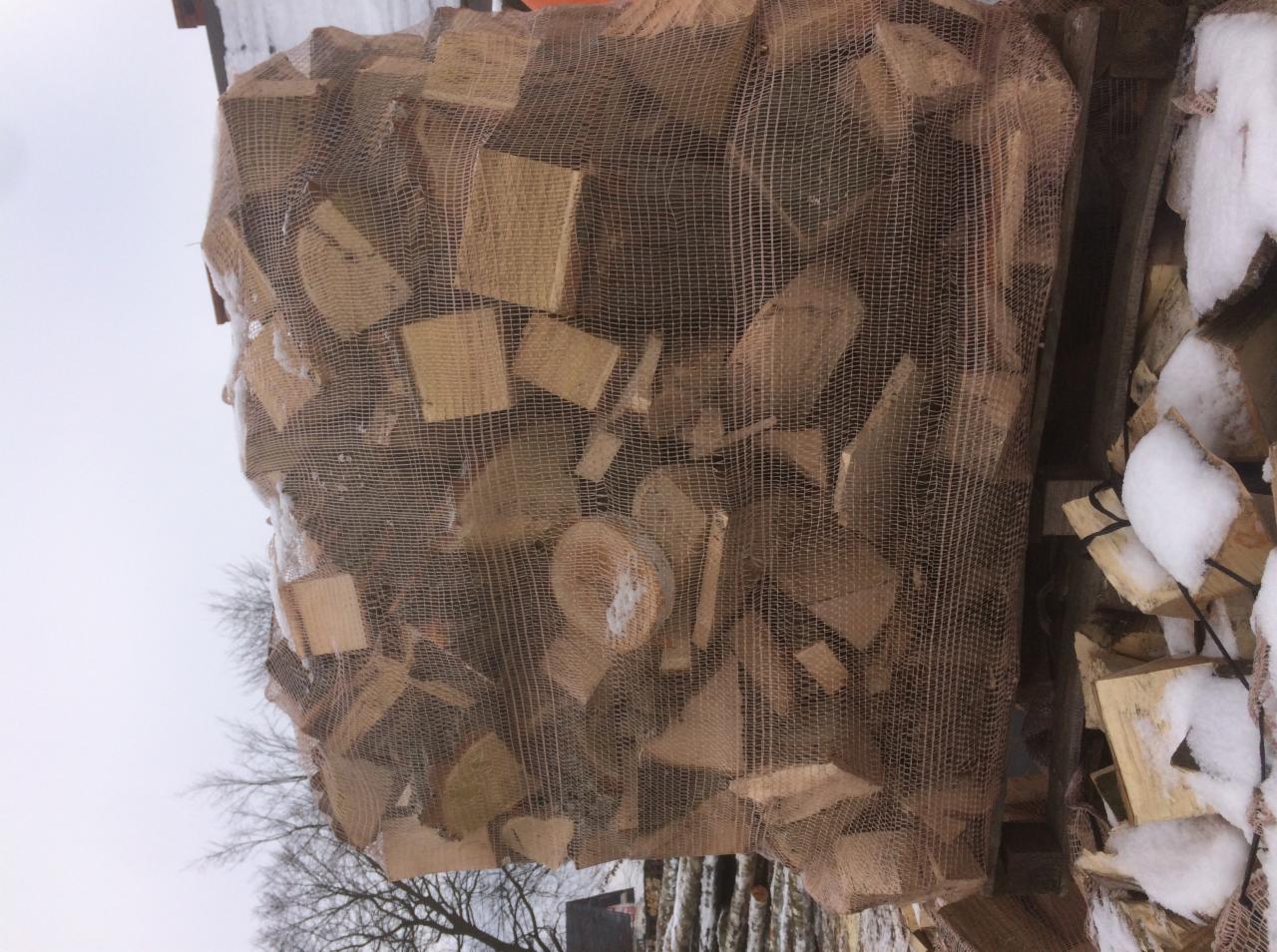 Siatka wypełniona pociętym drewnem ułożona na stosie drewna pokrytym śniegiem, widok z dołu na tle pochmurnego nieba i drzew bez liści.