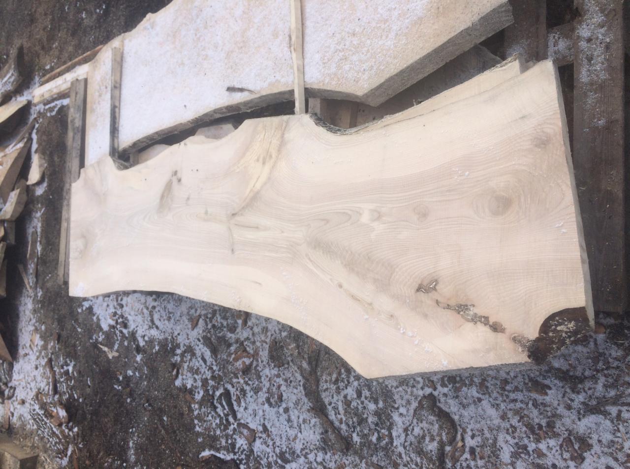 Surowe deski z jasnego drewna, ułożone na palecie na zewnątrz, widoczne sęki i naturalne krawędzie, lekko ośnieżone podłoże.