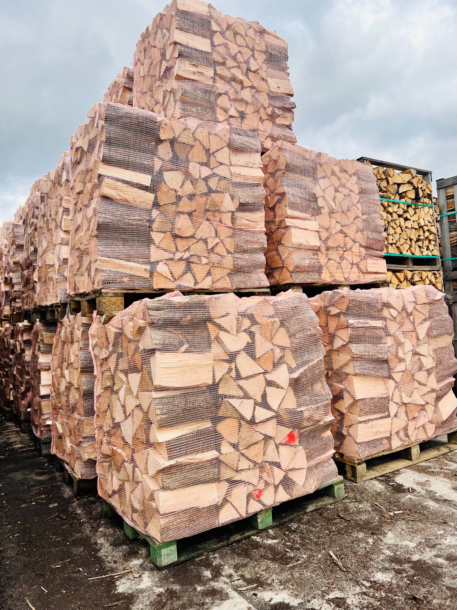 Stosy workowanego, pociętego drewna ułożone na paletach, widoczne inne palety z drewnem w tle, pochmurne niebo.