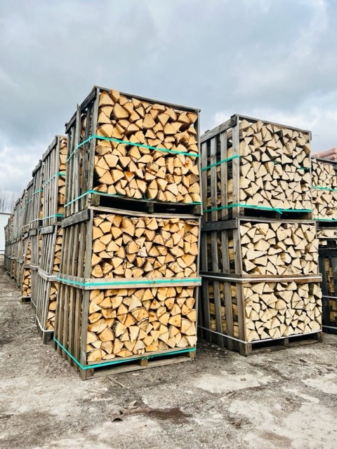 Ułożone w skrzyniach pocięte kawałki drewna opałowego, zabezpieczone zielonymi pasami, na zewnątrz pod pochmurnym niebem.