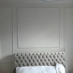 Jasnoszara tapicerowana rama łóżka z pikowaniem na tle białej ściany z dekoracyjnymi ramkami, widoczne gniazdka elektryczne.