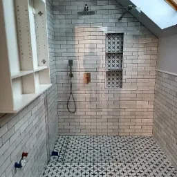 Nowoczesna kabina prysznicowa z białą cegiełką, czarno-białą mozaiką na podłodze i wnęką z dekoracyjnym wzorem, oświetlona światłem dziennym z okna dachowego.