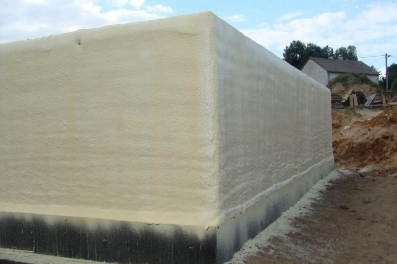 Ściana fundamentowa budynku ocieplona pianą PUR w kolorze kremowym, widoczne czarne ślady po izolacji bitumicznej, w tle dom jednorodzinny i plac budowy.