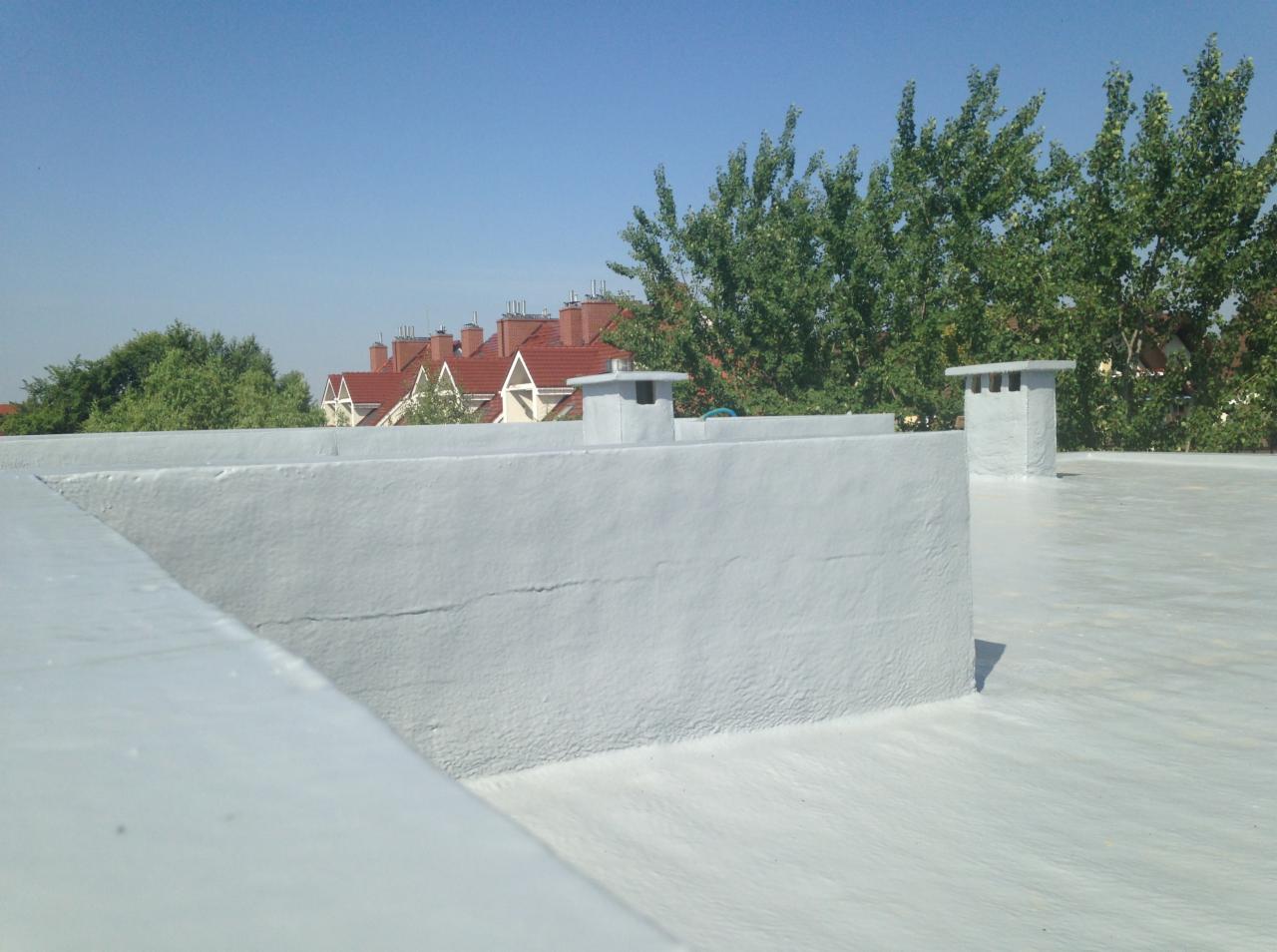 Płaski dach pokryty szarą, elastyczną powłoką hydroizolacyjną z widocznymi kominami wentylacyjnymi i budynkami w tle.