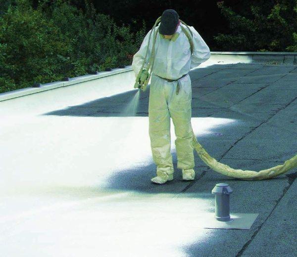 Pracownik w kombinezonie ochronnym natryskuje białą substancję na płaski dach pokryty papą, widoczny przewód doprowadzający materiał i kominek wentylacyjny.