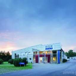 Budynek firmy HSK LEDY oświetlony ciepłym światłem zachodzącego słońca, z widocznym parkingiem i zielenią.