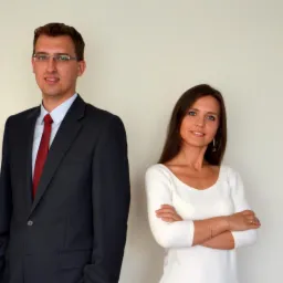 Portret dwojga prawników: mężczyzna w ciemnym garniturze i czerwonym krawacie, kobieta w białej bluzce z rękawami 3/4, oboje stoją na jasnym tle.