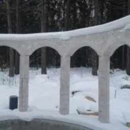 Niedokończona konstrukcja z kolumnami i łukami, prawdopodobnie altana lub element dekoracyjny ogrodu, pokryta śniegiem, w tle zimowy las.