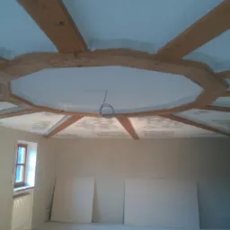 Wnętrze pokoju w trakcie prac wykończeniowych, widoczne drewniane belki stropowe w układzie rozchodzącym się od środka sufitu, częściowo pomalowany sufit i ściany, dwie płyty gipsowe oparte...