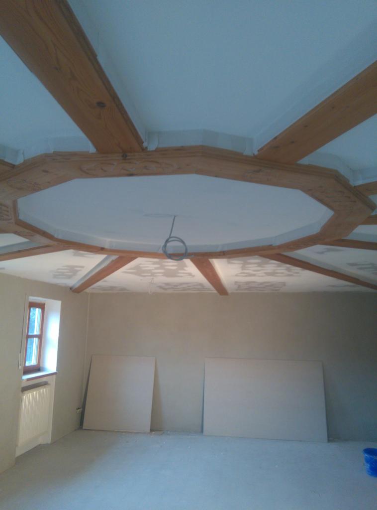 Wnętrze pokoju w trakcie prac wykończeniowych, widoczne drewniane belki stropowe w układzie rozchodzącym się od środka sufitu, częściowo pomalowany sufit i ściany, dwie płyty gipsowe oparte...