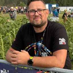 Uśmiechnięty mężczyzna w okularach, oparty o barierkę z napisem na tle tłumu i zieleni. Widoczny zegarek i tatuaż na ramieniu, koszulka z logo. Festiwal w tle.