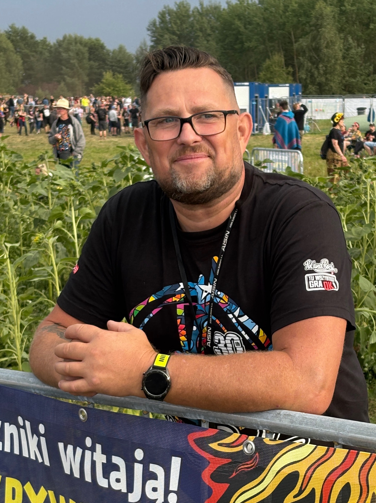 Uśmiechnięty mężczyzna w okularach, oparty o barierkę z napisem na tle tłumu i zieleni. Widoczny zegarek i tatuaż na ramieniu, koszulka z logo. Festiwal w tle.