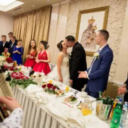 Nowożeńcy całują się za stołem weselnym udekorowanym kwiatami i jedzeniem, w tle goście i obraz Matki Boskiej Częstochowskiej.