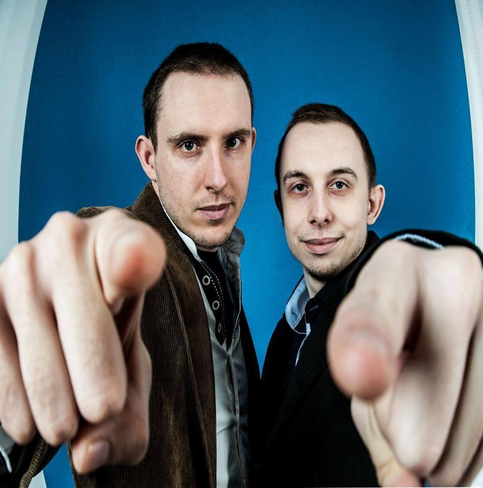 Portret dwóch mężczyzn wskazujących palcami w kierunku widza na tle niebieskiego tła, z efektem rybiego oka.