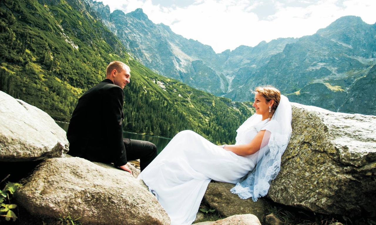 Para młoda siedzi na kamieniach z widokiem na góry; panna młoda w białej sukni i welonie, pan młody w czarnym garniturze.