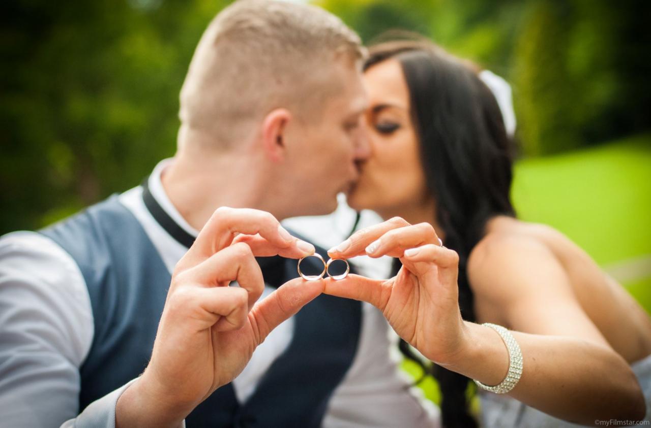 Pocałunek pary młodej z obrączkami ślubnymi prezentowanymi na dłoniach na tle zieleni.