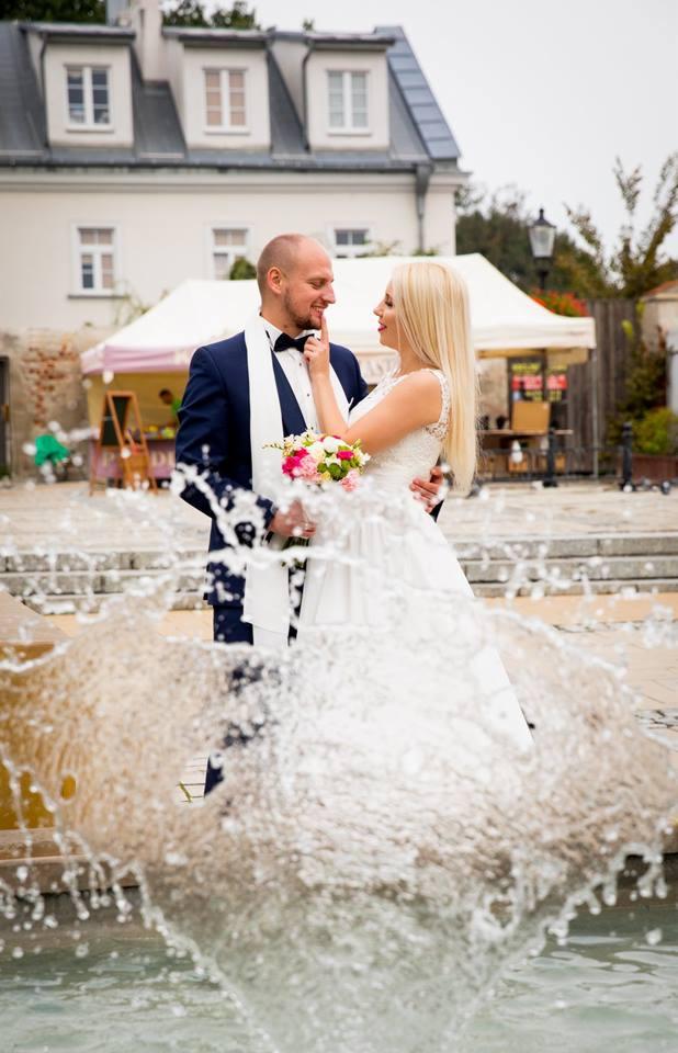 Uśmiechnięta para młoda, panna młoda dotyka brody pana młodego, na tle fontanny z rozpryskującą się wodą i budynkiem w tle.