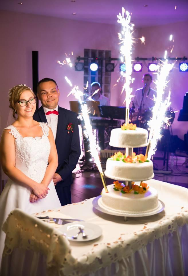 Para młoda uśmiechnięta, obok trzypiętrowy tort weselny z zimnymi ogniami, w tle zespół muzyczny na scenie.